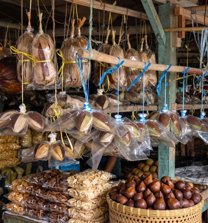 Gambar mendetail dari stan pasar tradisional Indonesia yang padat dan autentik, mendemonstrasikan visual merchandising pasar tradisional yang cerdas. Di bagian atas, logo emas 'gula aren cetak' dipajang di struktur kayu. Di bawahnya, banyak kemasan plastik berisi gula aren organik yang digantung dari tiang bambu dengan tali rafia biru dan kuning. Di meja kayu depan, keripik renyah dikemas dalam tumpukan plastik, dan keranjang anyaman besar penuh dengan buah salak di sebelah kanan. Gambar ini mengilustrasikan strategi display produk umkm yang efektif untuk meningkatkan nilai jual produk lokal.