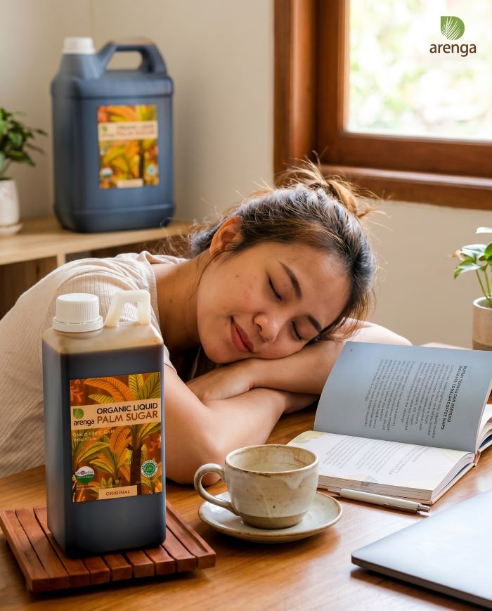 Seorang wanita muda tertidur sejenak di atas meja kayu dengan kepala bertumpu pada lengannya. Di depannya terdapat cangkir kopi kosong dan botol gula aren cair Arenga 1kg. Di latar belakang, terlihat jerigen besar gula aren Arenga 10 liter di atas rak dan sebuah buku yang terbuka. Cahaya alami masuk dari jendela. Logo Arenga terlihat di sudut kanan atas.