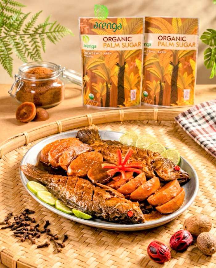 Foto hidangan Semur Ikan Manado yang menggugah selera, menampilkan dua ekor ikan goreng utuh dengan kuah semur kental dan potongan kentang di atas nampan bambu anyaman. Di latar belakang terdapat dua kemasan standing pouch Arenga Organic Palm Sugar (Gula Semut Aren), toples gula, serta rempah-rempah utuh seperti cengkeh dan pala.